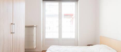 A bedroom with a bed, a wardrobe, a radiator, and a window with a view of buildings.