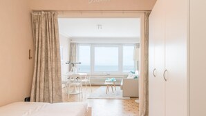 A bedroom with a bed, a desk, a chair, and a view of the sea through large windows. - Frontal sea View Studio Apartment in Ostend (Ostend)
