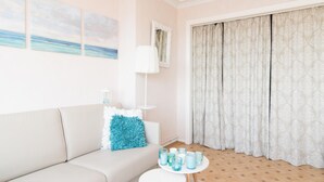 A modern living room with a beige sofa, a white coffee table, and a lamp. There are three paintings on the wall, a window with curtains, and a patterned carpet. - Frontal sea View Studio Apartment in Ostend (Ostend)