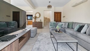A modern living room with a grey sectional sofa, a coffee table, and a TV on a wooden stand. There is a staircase leading to another level, and a wooden door on the right.