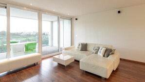 A modern living room with a white sofa, a white coffee table, and a bench. There are large windows providing a view of the city. - South Facing Apartment With Terrace (Ostend)