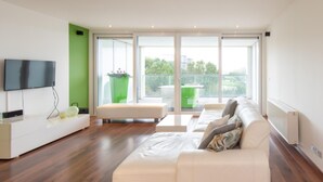 A modern living room with a large white sectional sofa, a flat-screen TV mounted on the wall, and a balcony with a green planter. - South Facing Apartment With Terrace (Ostend)