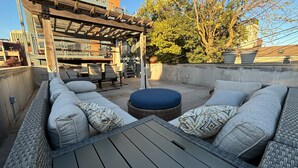A rooftop patio with a wicker sofa set, a round ottoman, and a pergola structure.