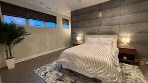A modern bedroom with a large bed, two bedside lamps, a potted plant, and a view of the sky through the windows.