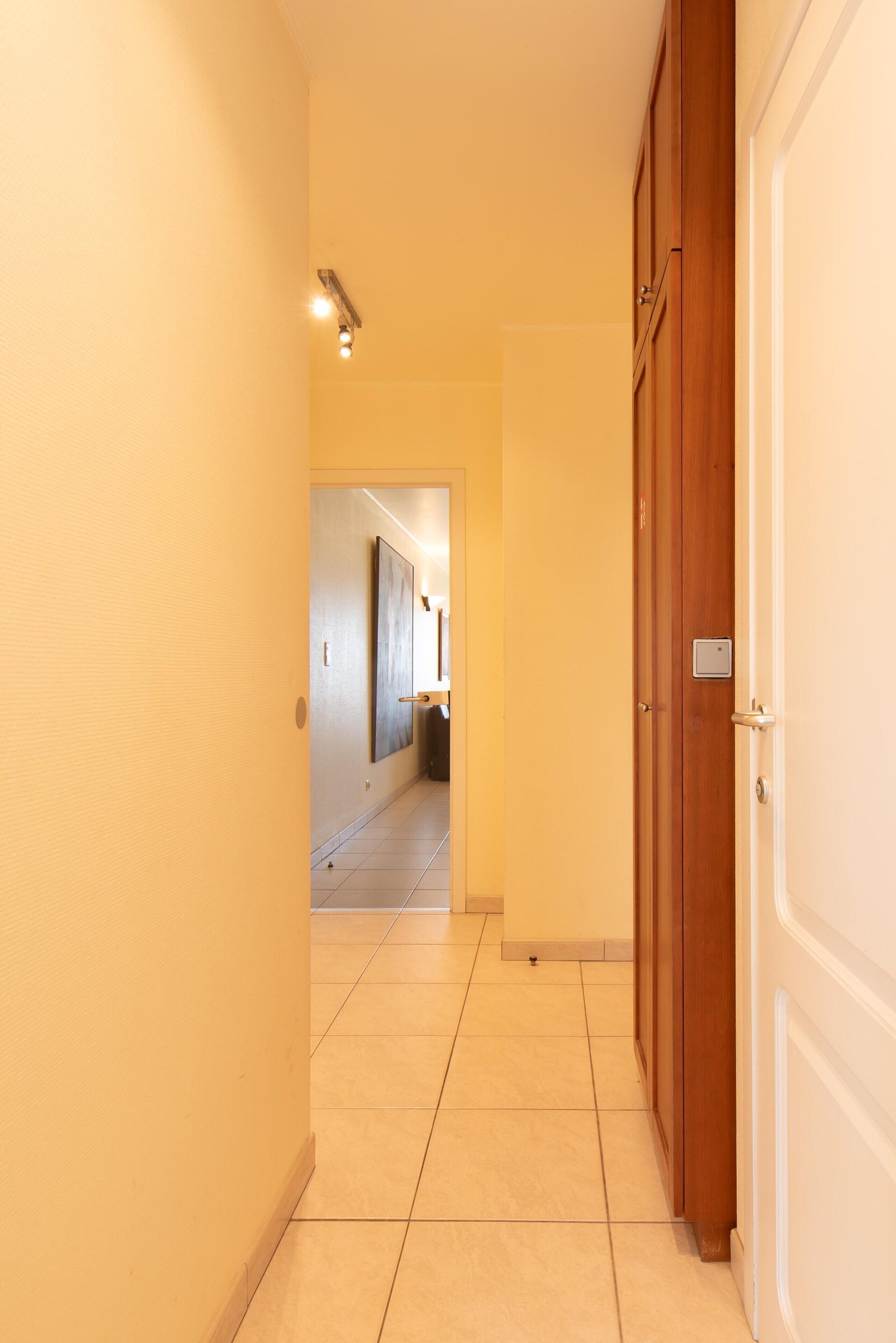 Un corridoio con una porta in legno sulla destra, un lampadario a sinistra e un pavimento piastrellato.