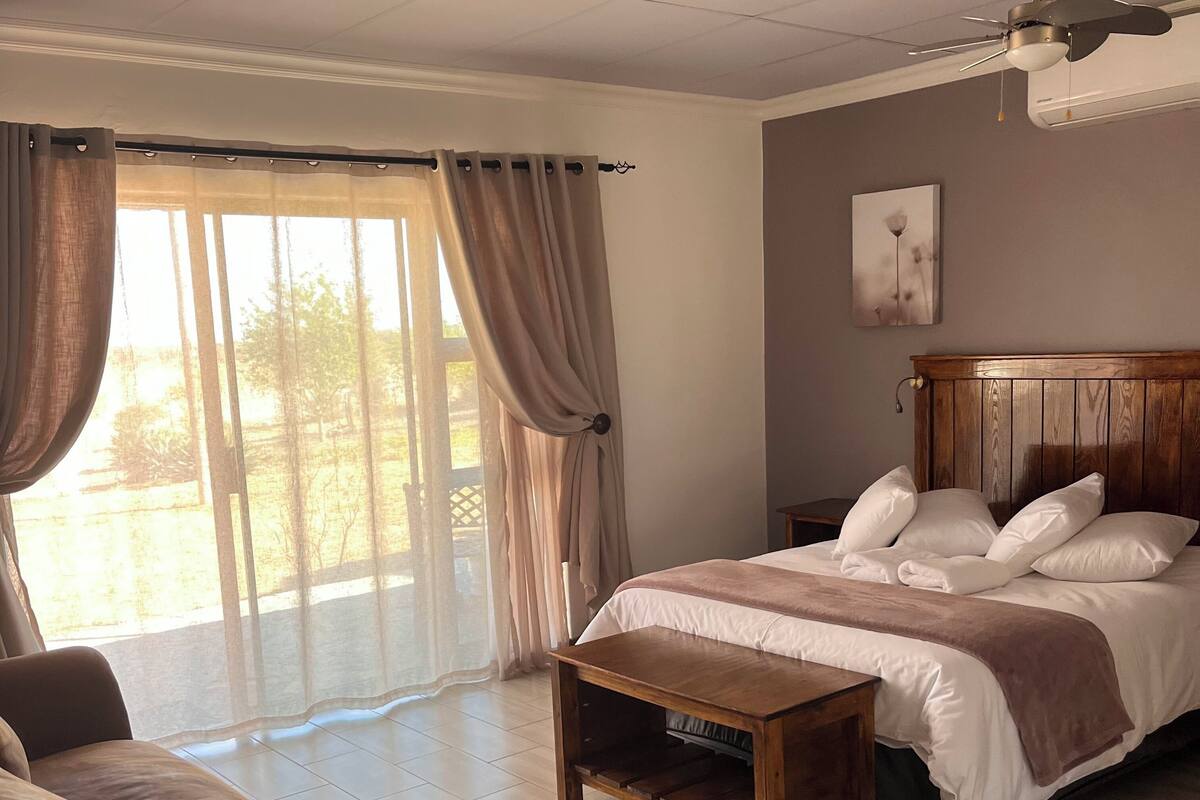 Un dormitorio con cama, cabecera de madera, mesita de noche y vistas a través de un gran ventanal con cortinas.
