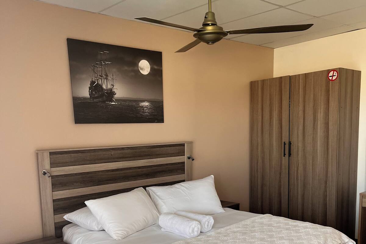 Un dormitorio con cama, mesitas de noche, ventilador de techo, armario y un cuadro en la pared de un barco en el mar.