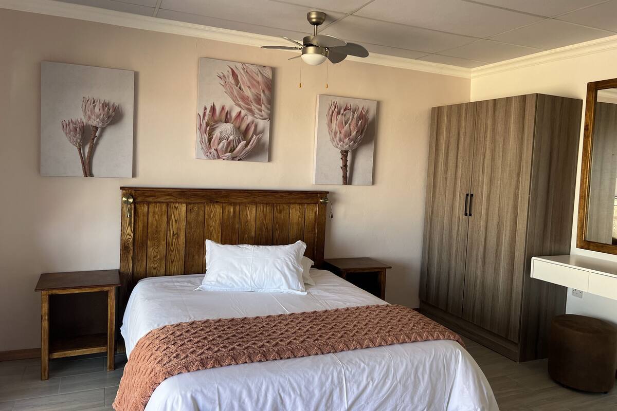 Un dormitorio con cabecera de madera, una cama grande con ropa de cama blanca, un plaid tejido marrón y tres cuadros colgados en la pared que representan flores.
