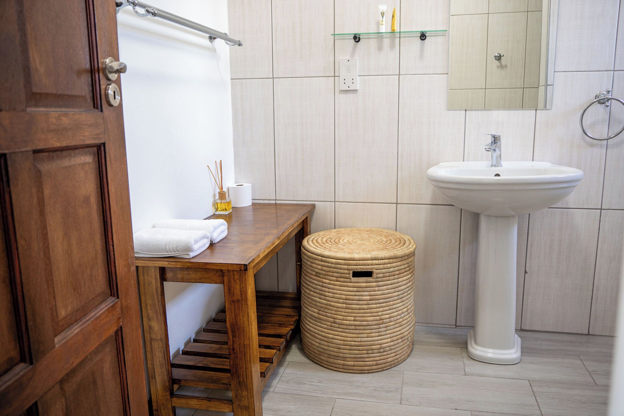Une salle de bain avec un banc en bois, un panier rond en osier, un lavabo blanc et un miroir au-dessus du lavabo.