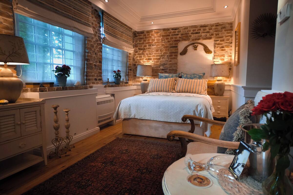 A bedroom with a brick wall, a large window, a bed with striped pillows, a bedside table with a lamp, and a vase with flowers.