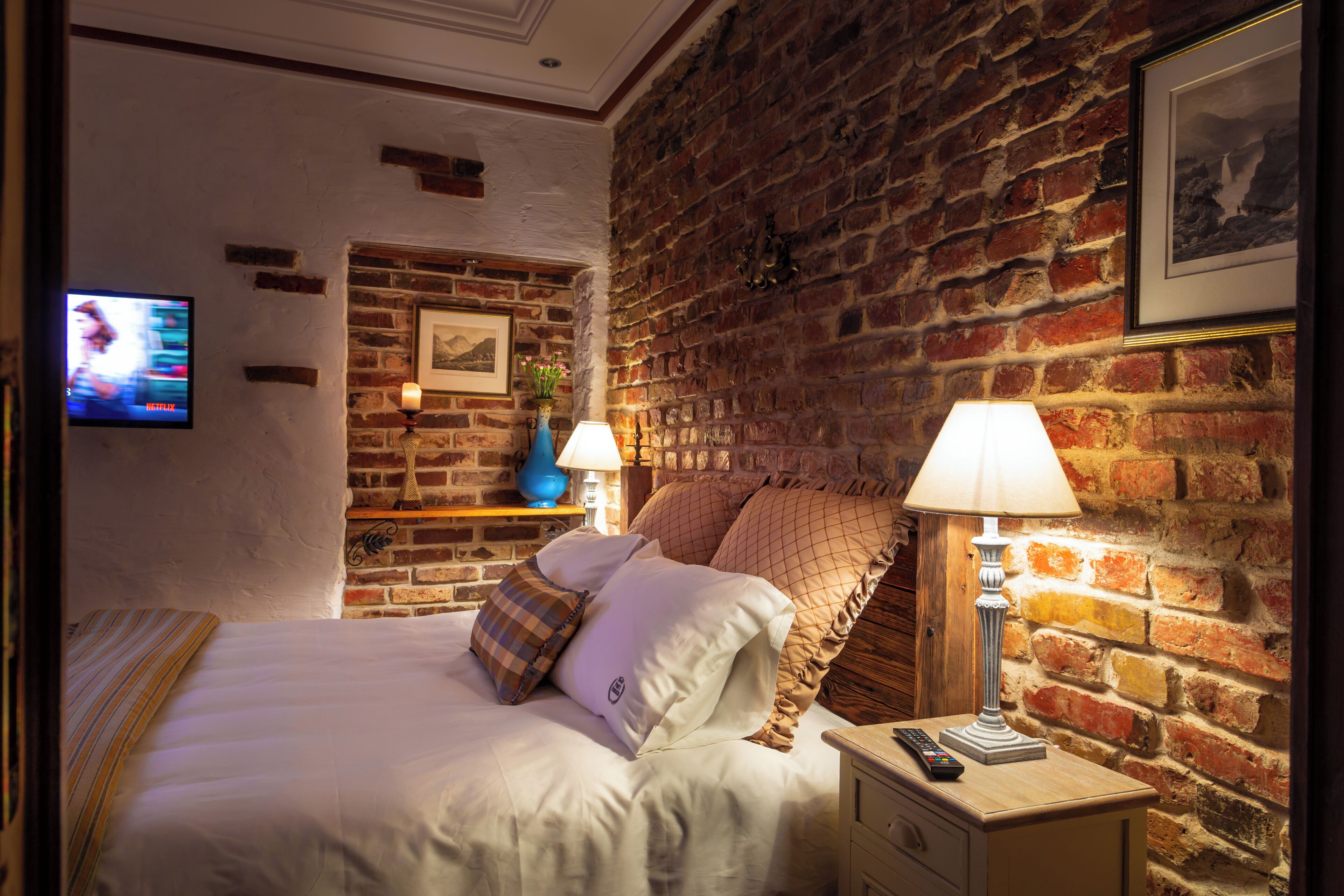 A hotel room with a brick wall, a bed with white bedding, a nightstand with a lamp, and a TV on the wall.