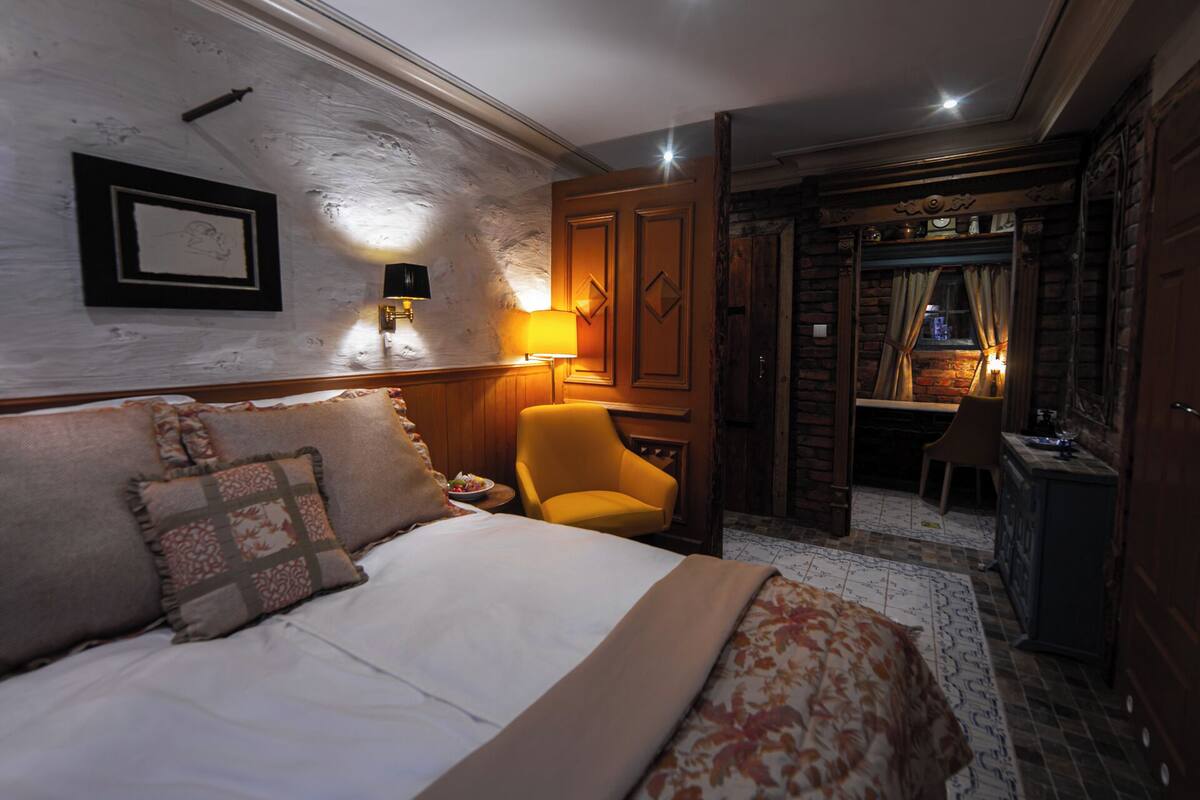 A hotel room with a bed, a chair, a desk, and a view of a dining area.