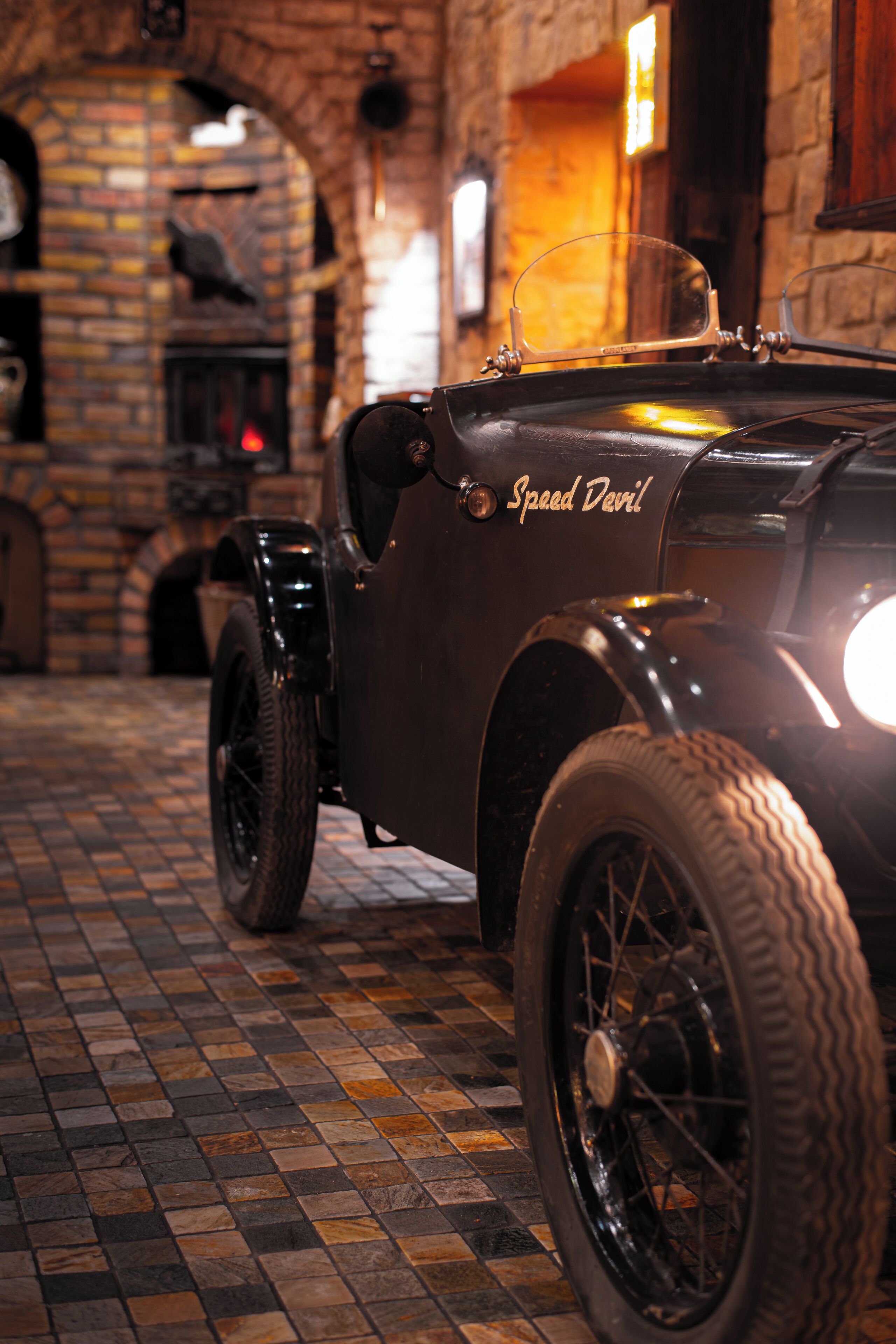 A vintage car with "Speed Devil" written on its side, parked indoors with a brick wall and a fireplace in the background.