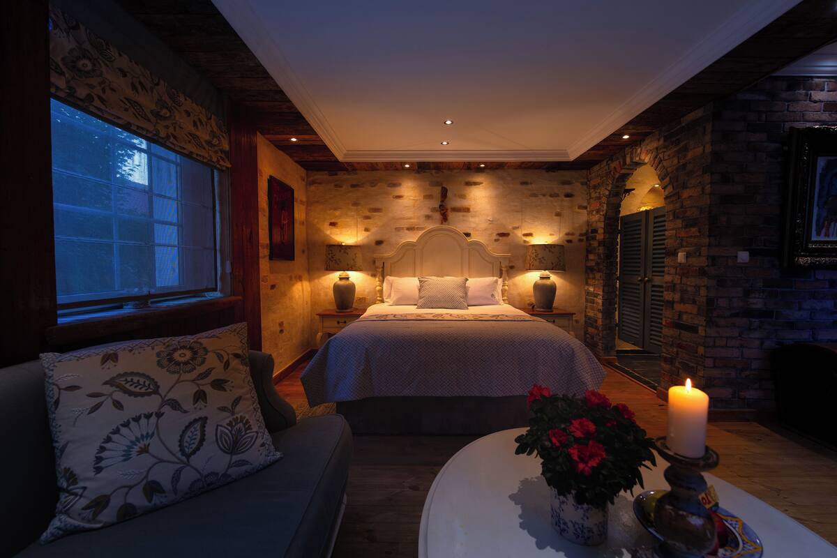 A cozy bedroom with a large bed, a sofa, a bedside table with a candle, and a window with blinds.