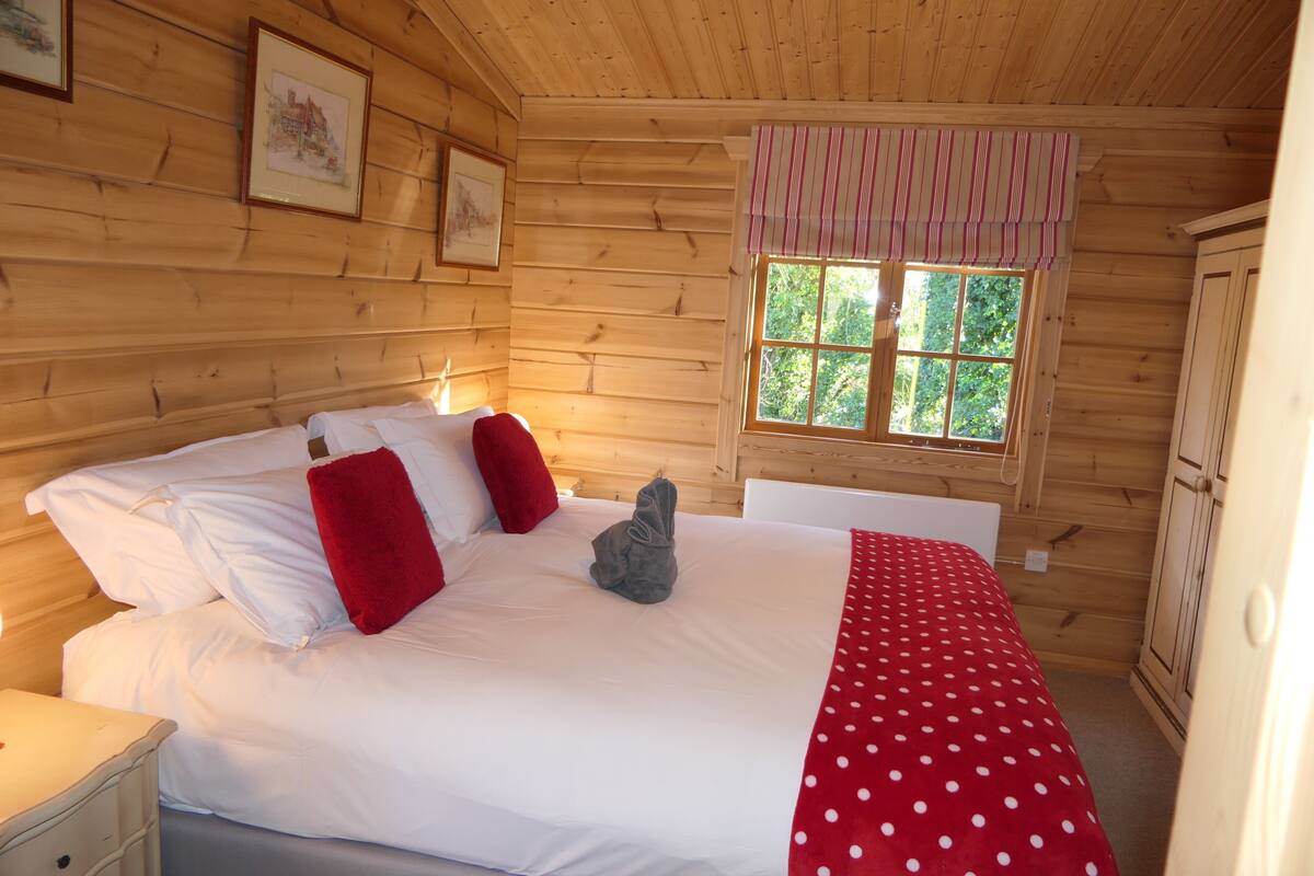 A wooden cabin bedroom with a bed, pillows, a red polka dot blanket, a window with a striped valance, and wooden walls.