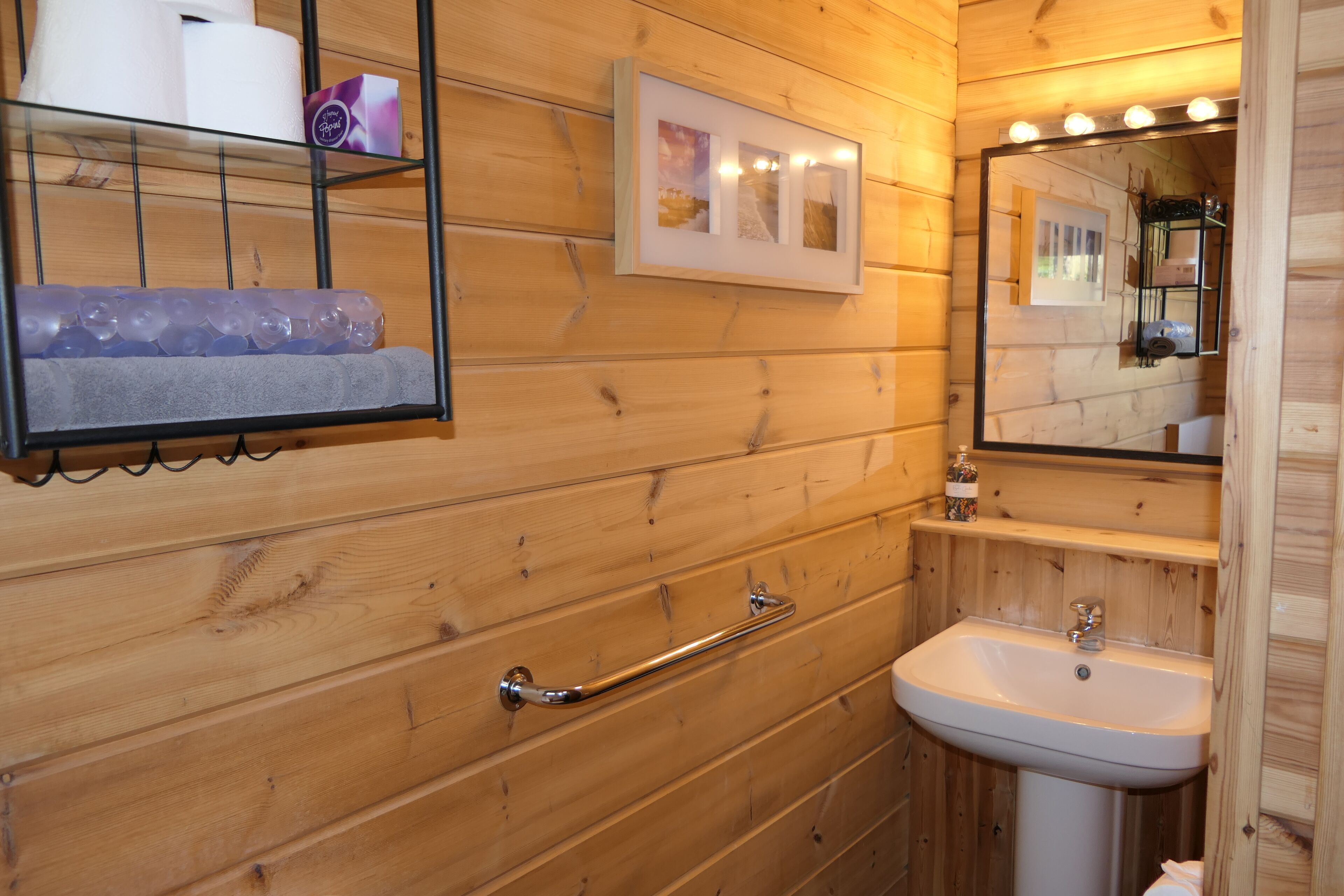 Ein Badezimmer mit Holzpaneelen, einem weißen Waschbecken, einer Handstange, einem Ablagefach mit Pflegeprodukten und einem gerahmten Bild an der Wand.
