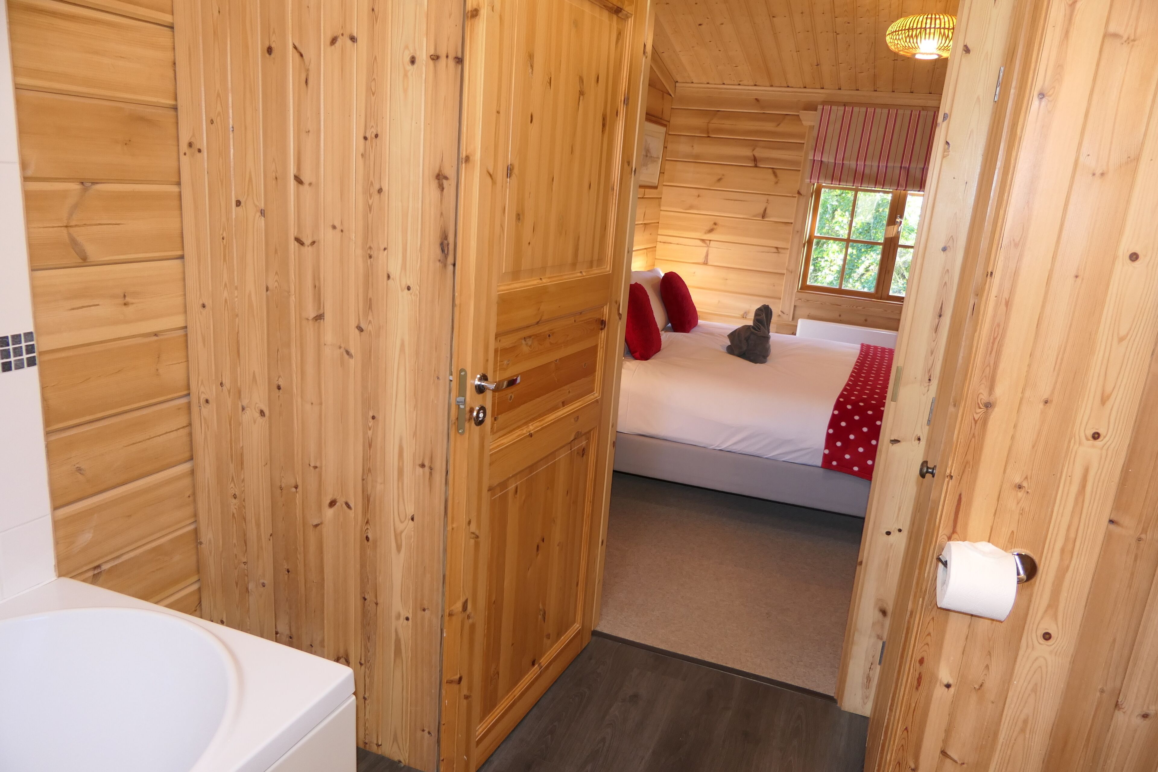 A wooden cabin room with a bed, a bathroom with a white bathtub, and a window with a red curtain.