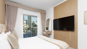 A hotel room with a large bed, a TV mounted on the wall, a desk with a chair, and a view of the city through the window.