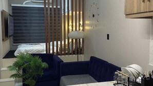 A modern hotel room with a sofa, a small table with a plant, a desk with a lamp, and a view of the bed and window blinds. -  smart, affordable condo unique blend of luxury, convenience, and modern living (Quezon City)
