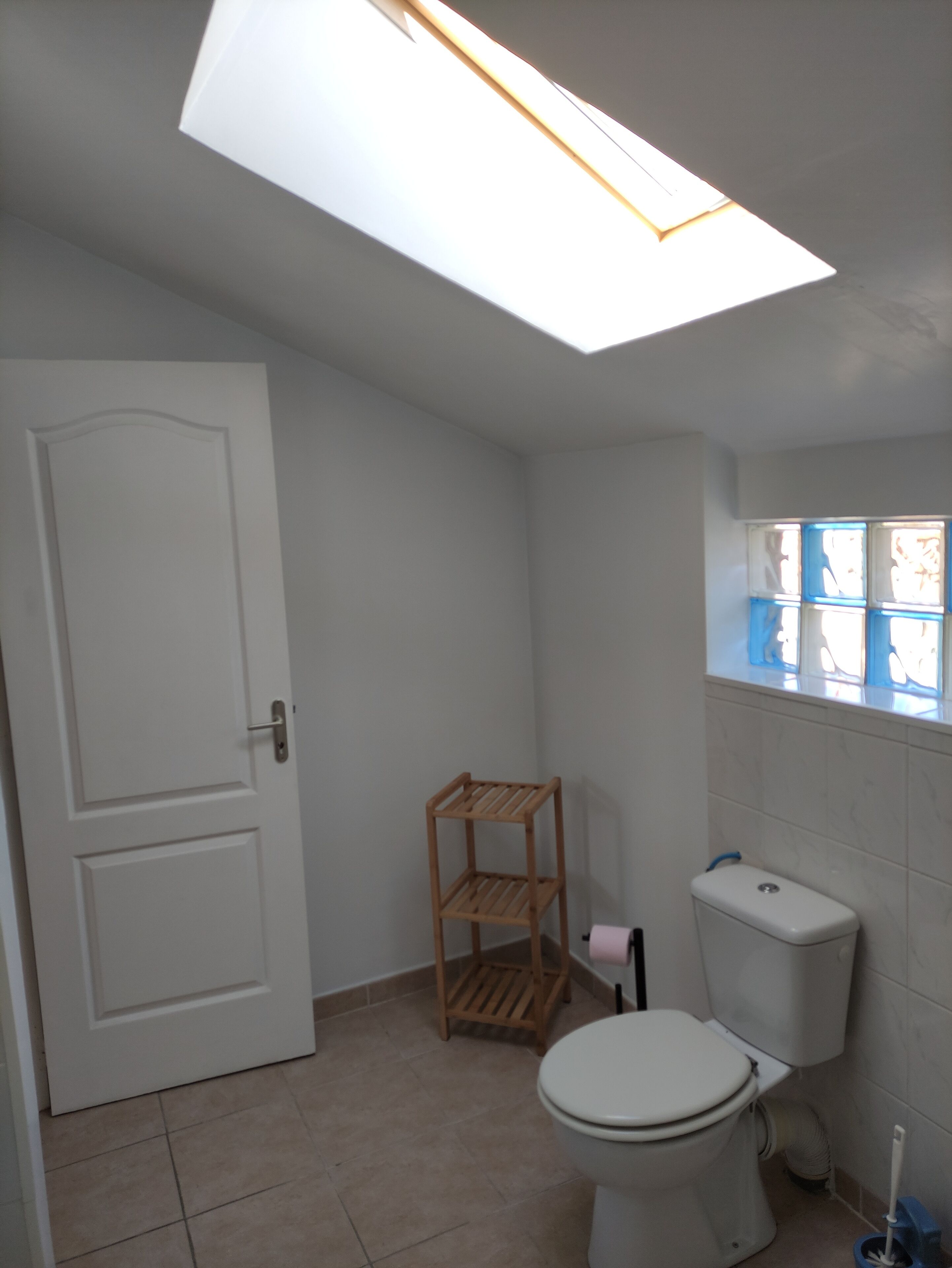 Ein Badezimmer mit Oberlicht, Toilette, Holzregal und Fenster mit blauen Rahmen.
