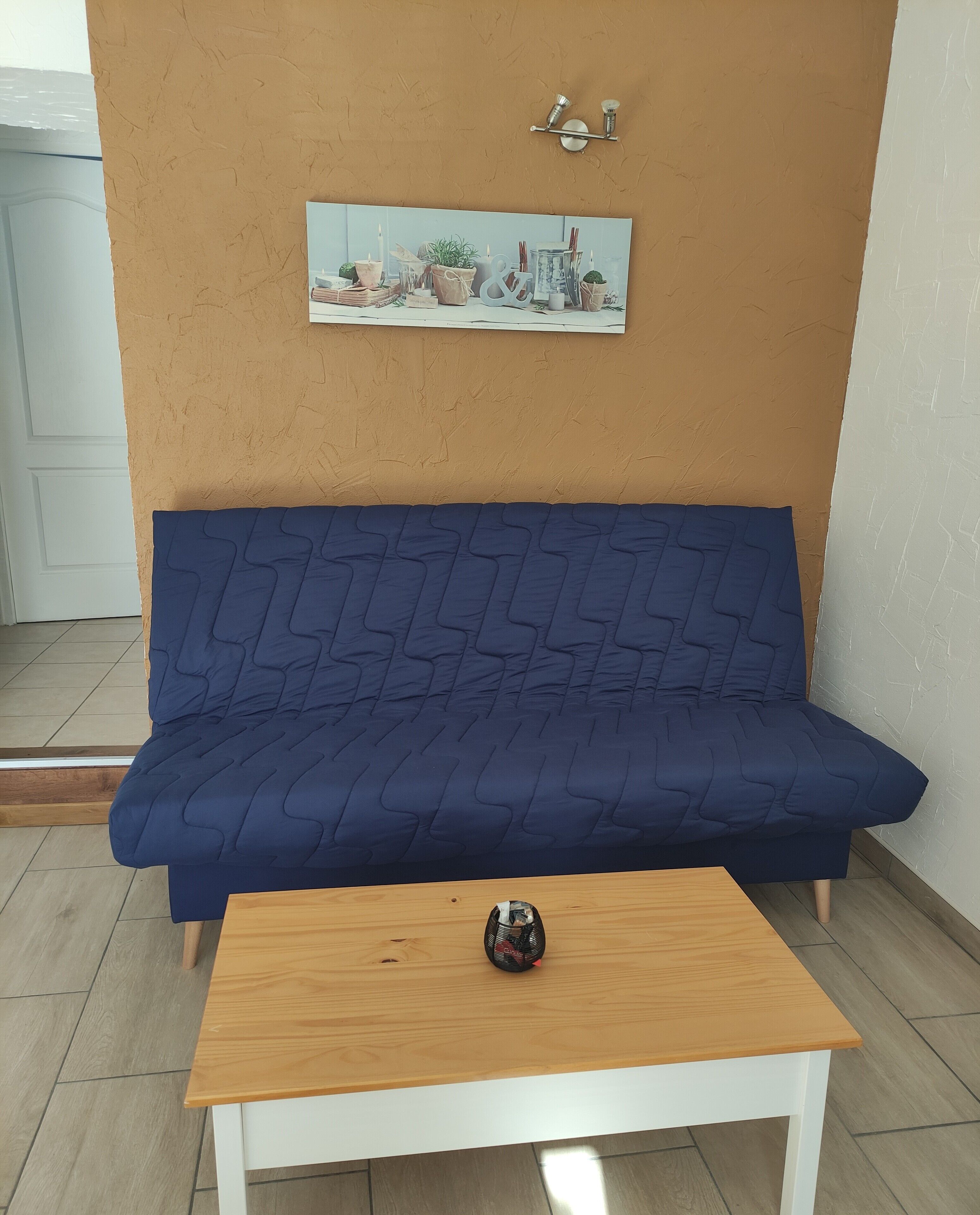 Ein blaues Sofa mit strukturierter Rückenlehne, ein Couchtisch aus Holz mit einem dekorativen Gegenstand und ein wandmontiertes Regal mit einem gerahmten Bild und zwei Wandleuchten.