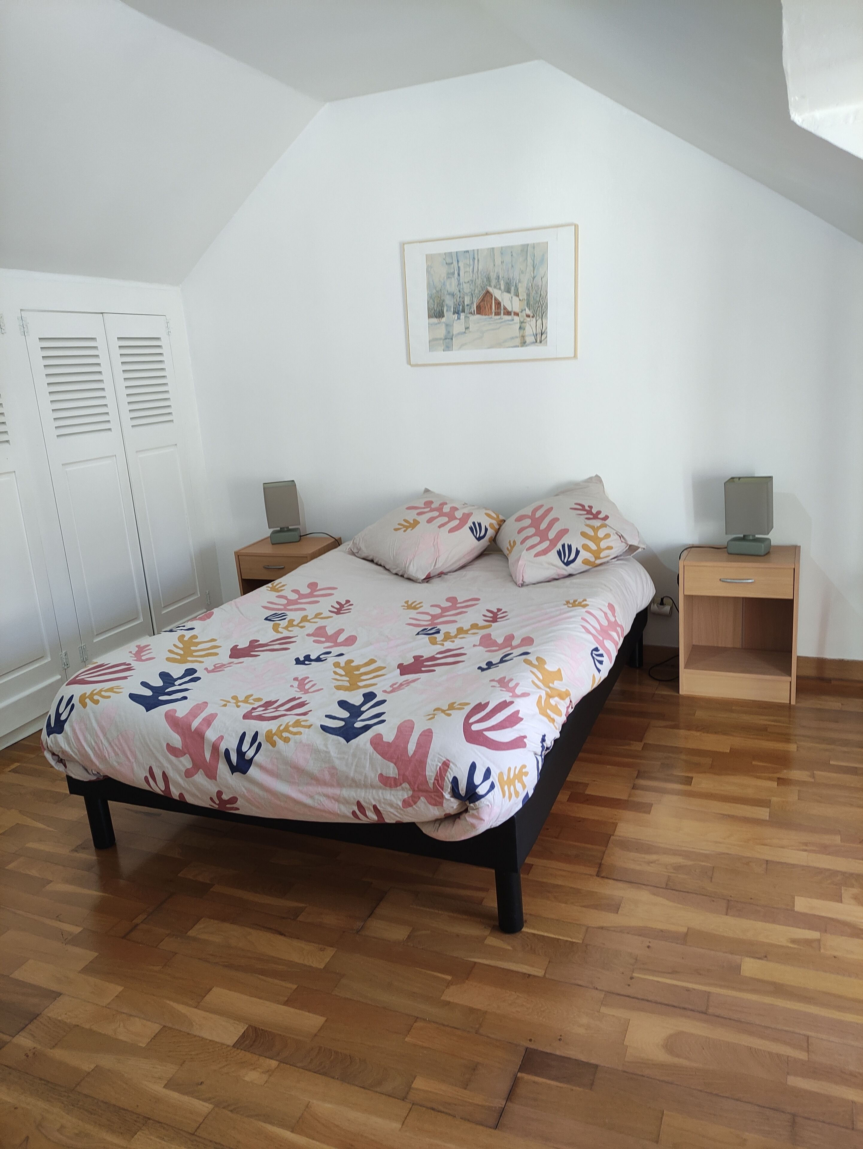 Une chambre à coucher comprenant un lit, deux tables de chevet, une commode, un tableau encadré au mur et un placard avec des portes à persiennes.