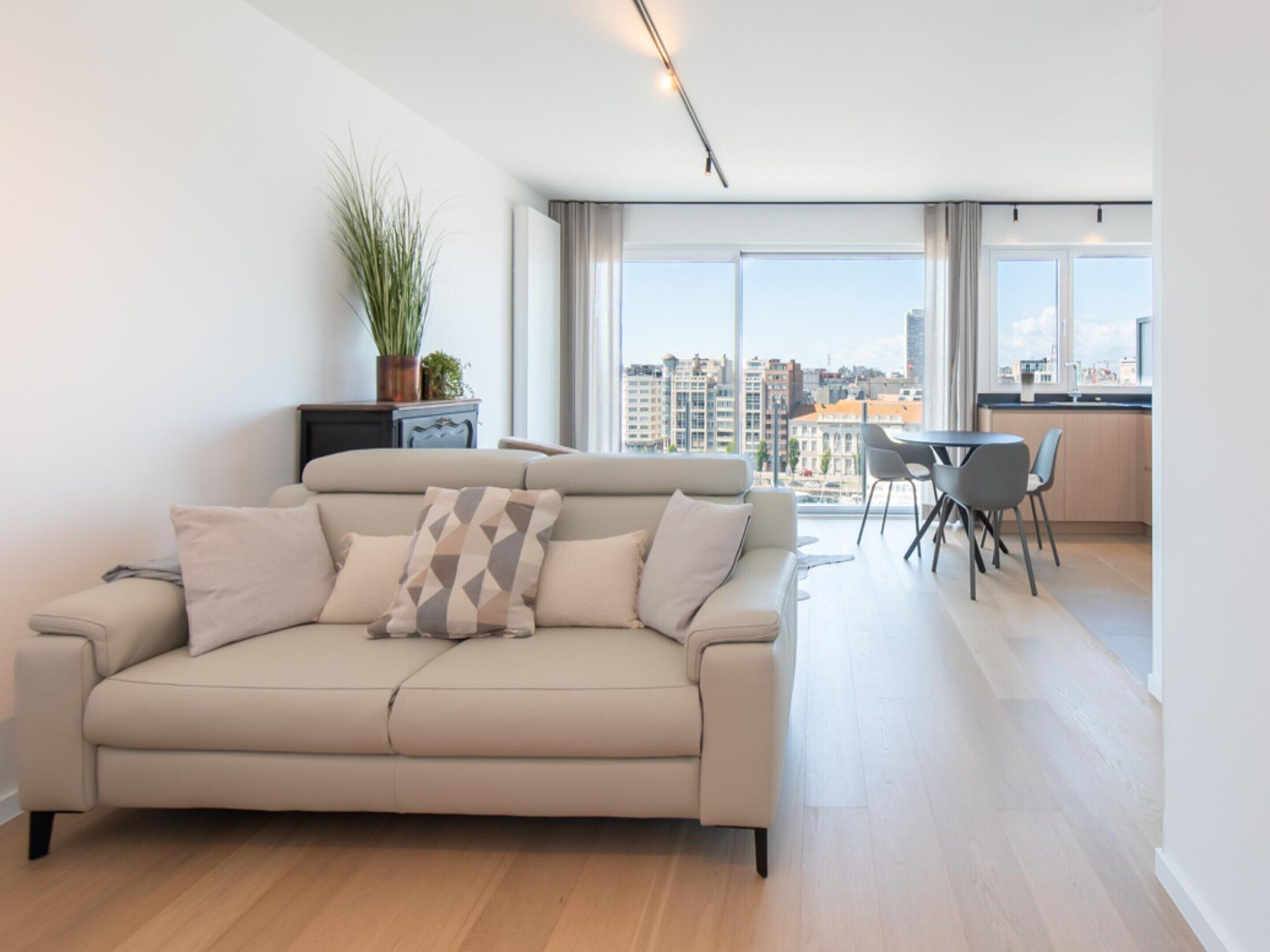 Un salon moderne avec un canapé beige, un coin repas comprenant une table et des chaises, et de grandes fenêtres offrant une vue sur la ville.