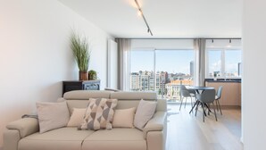 A modern living room with a beige sofa, a dining area with a table and chairs, and large windows offering a city view. - Apartment in Ostend Near Marina & Park (Ostend)