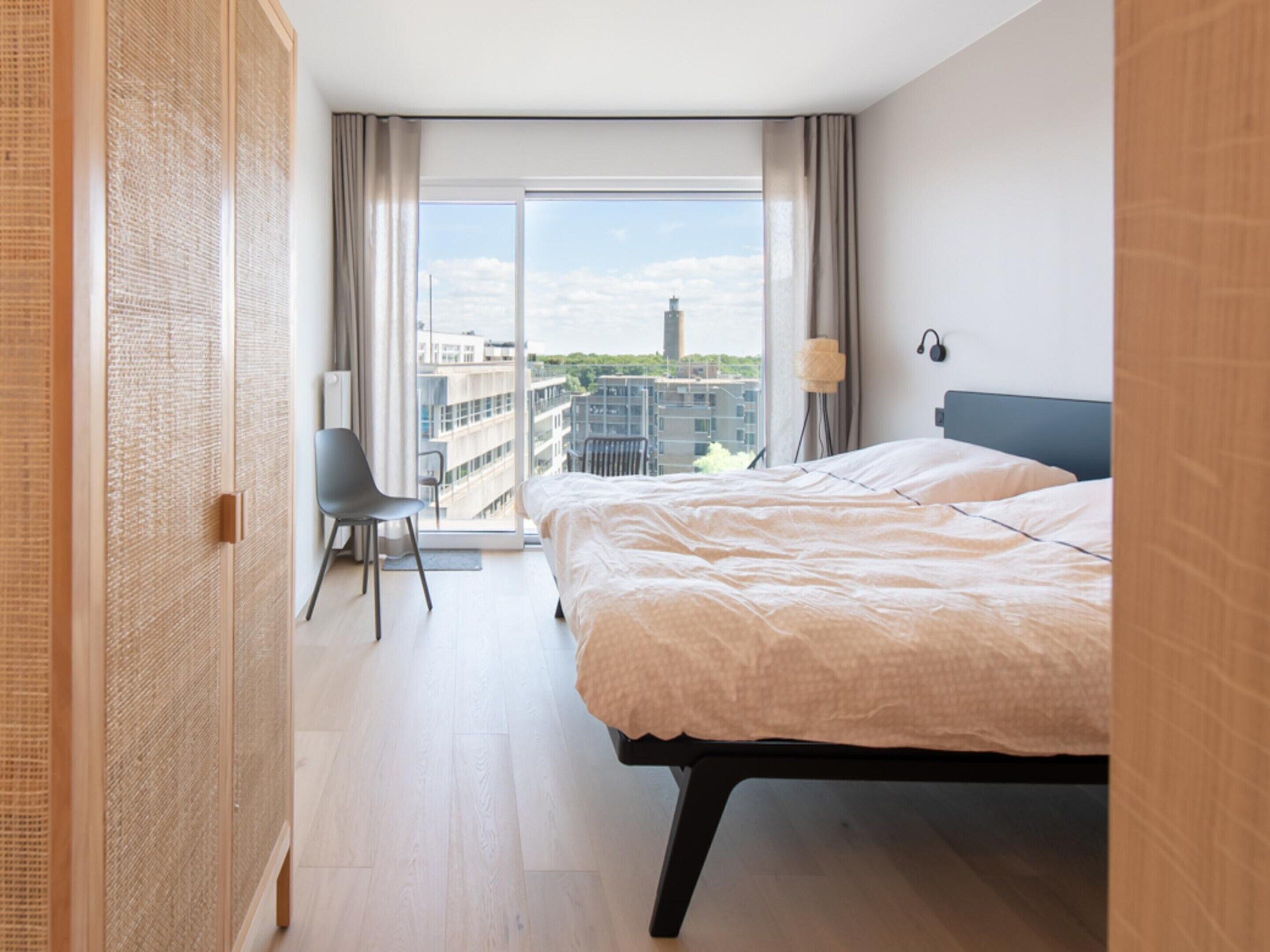 A modern hotel room with a large bed, a chair, a desk, and a view of the cityscape through a large window.