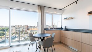 A modern kitchen with a large window, a round dining table, and a view of the city. - Apartment in Ostend Near Marina & Park (Ostend)