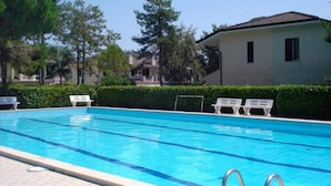 A swimming pool with clear blue water, surrounded by white lounge chairs and a hedge. - Holiday Home in Porto Santa Margherita Beach (Caorle)