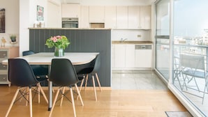 A modern kitchen with white cabinets, a dining area with a table and chairs, and a view through a large window. - Apartment in Ostend Near Shopping Street (Bons Villers)