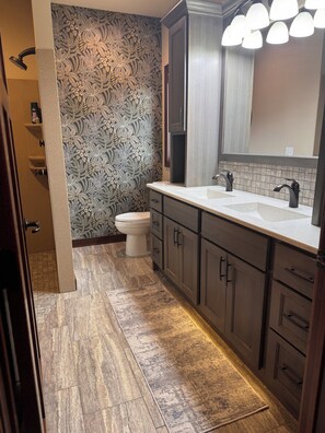 A bathroom with a double sink, dark wood cabinets, a patterned wallpaper, and a shower area.