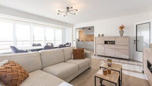 A modern living room with a sofa, coffee table, and dining area. A kitchen is visible in the background with wooden cabinets and a dining table. - Modern Flat Near the sea (Middelkerke)