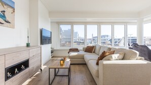 A modern living room with a sofa, coffee table, and TV. Large windows offer a city view. - Modern Flat Near the sea (Middelkerke)