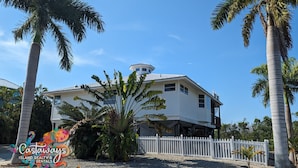 Ein weißes, zweistöckiges Haus mit weißem Zaun, umgeben von Palmen und blauem Himmel.