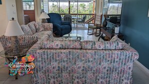 Ein Wohnzimmer mit einem blumigen Sofa, einem blauen Sessel, einem Glastisch und einem Fernseher.