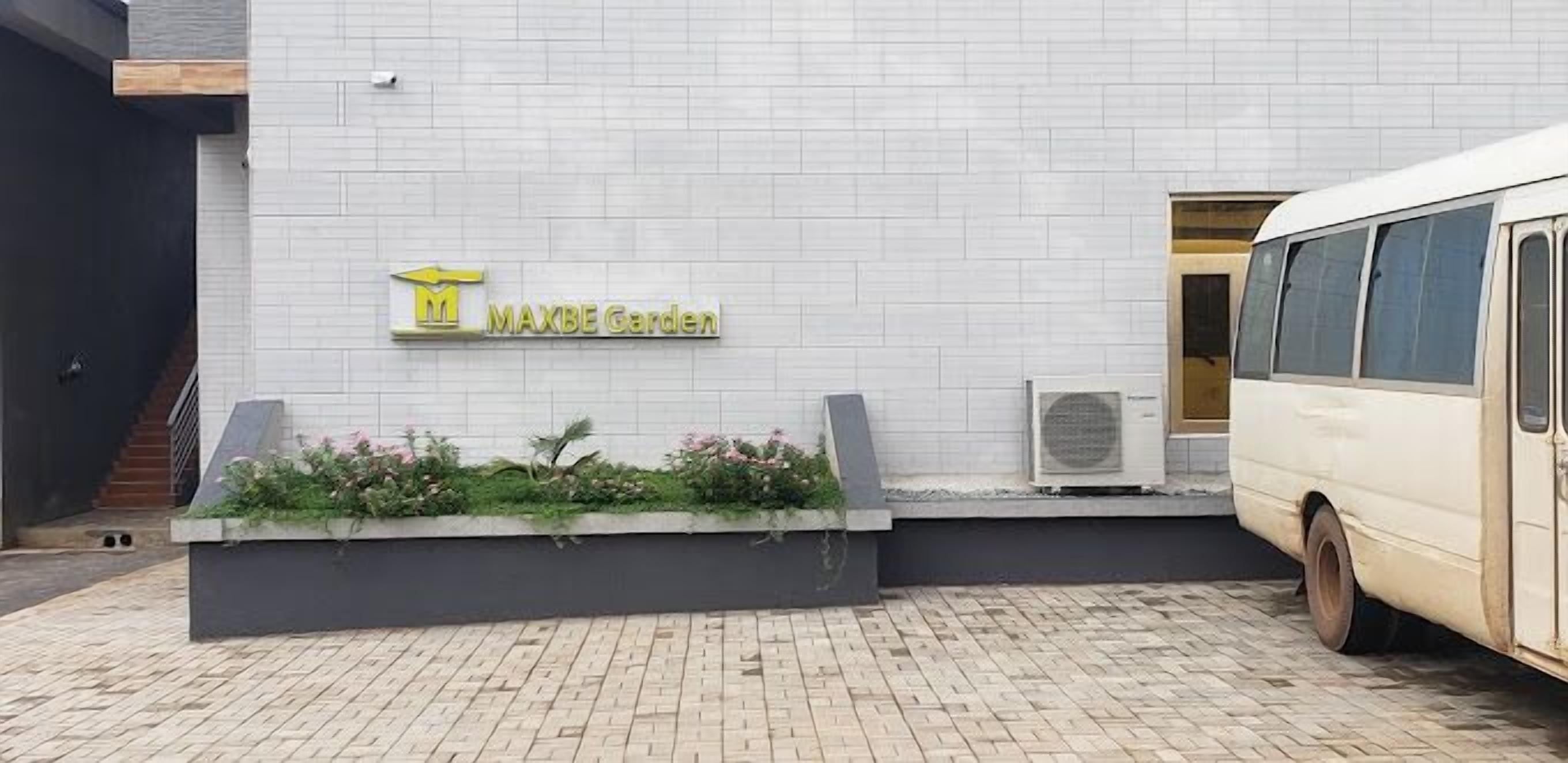 A MAXBE Garden sign on a white tiled wall, a bus parked to the right, and a planter with greenery in front.