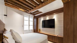 Une chambre moderne avec un grand lit, des poutres en bois au plafond, un téléviseur intégré et une fenêtre.