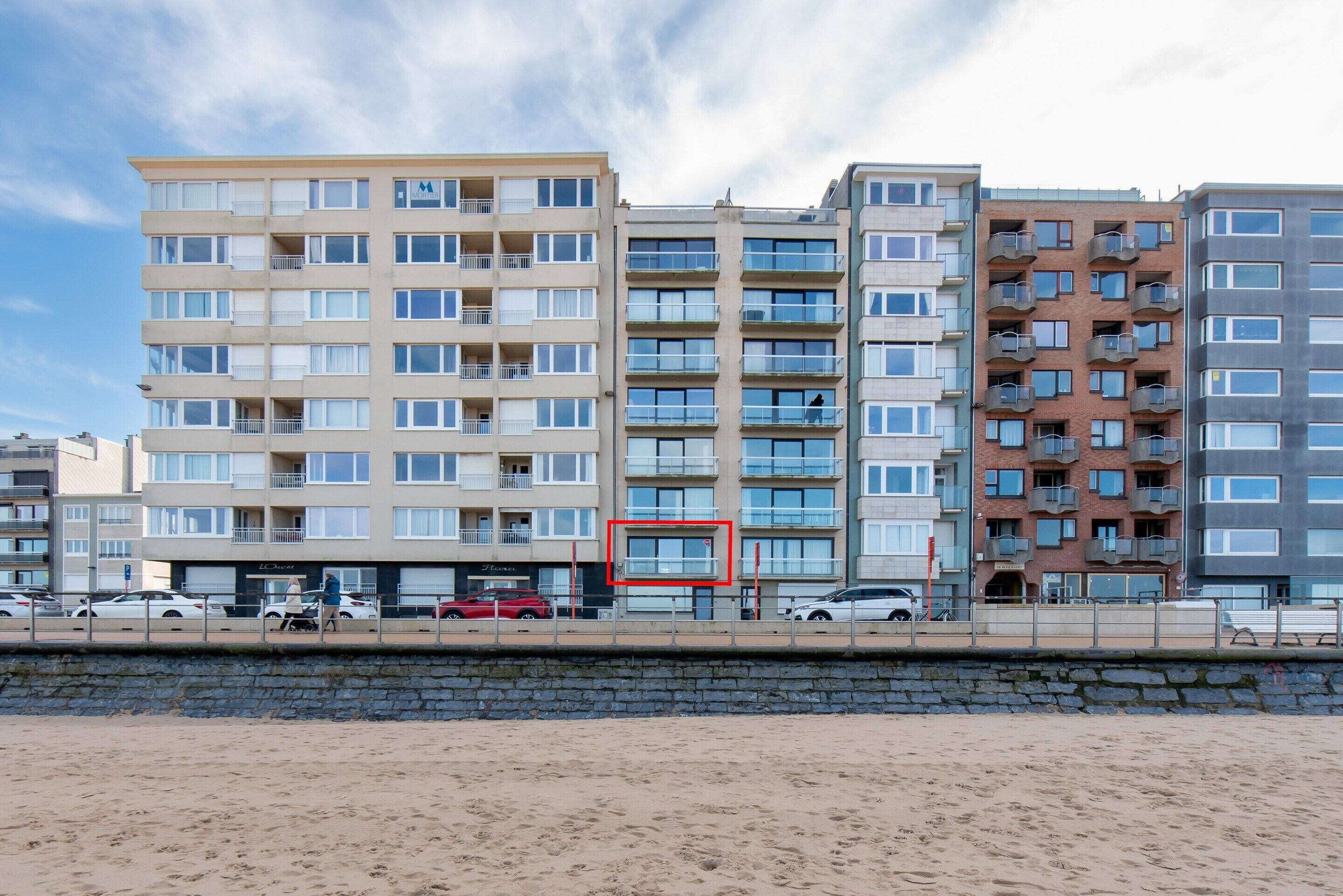 A multi-story residential building with balconies, a beach in the foreground, and a clear sky.