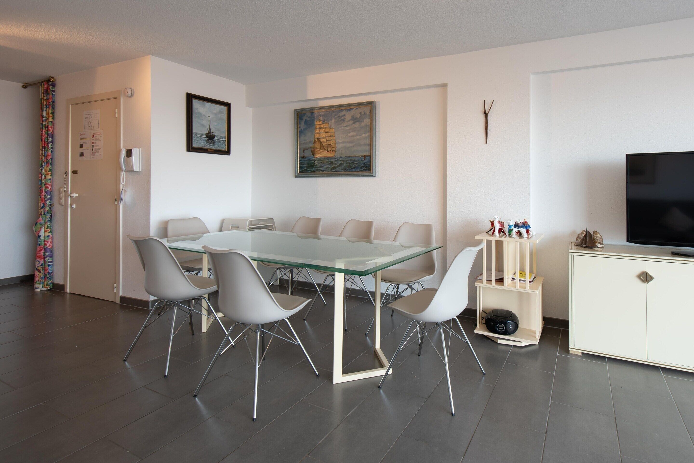 A modern living room with a glass-top dining table surrounded by white chairs, a flat-screen TV, a white cabinet, and a painting of a sailboat on the wall.