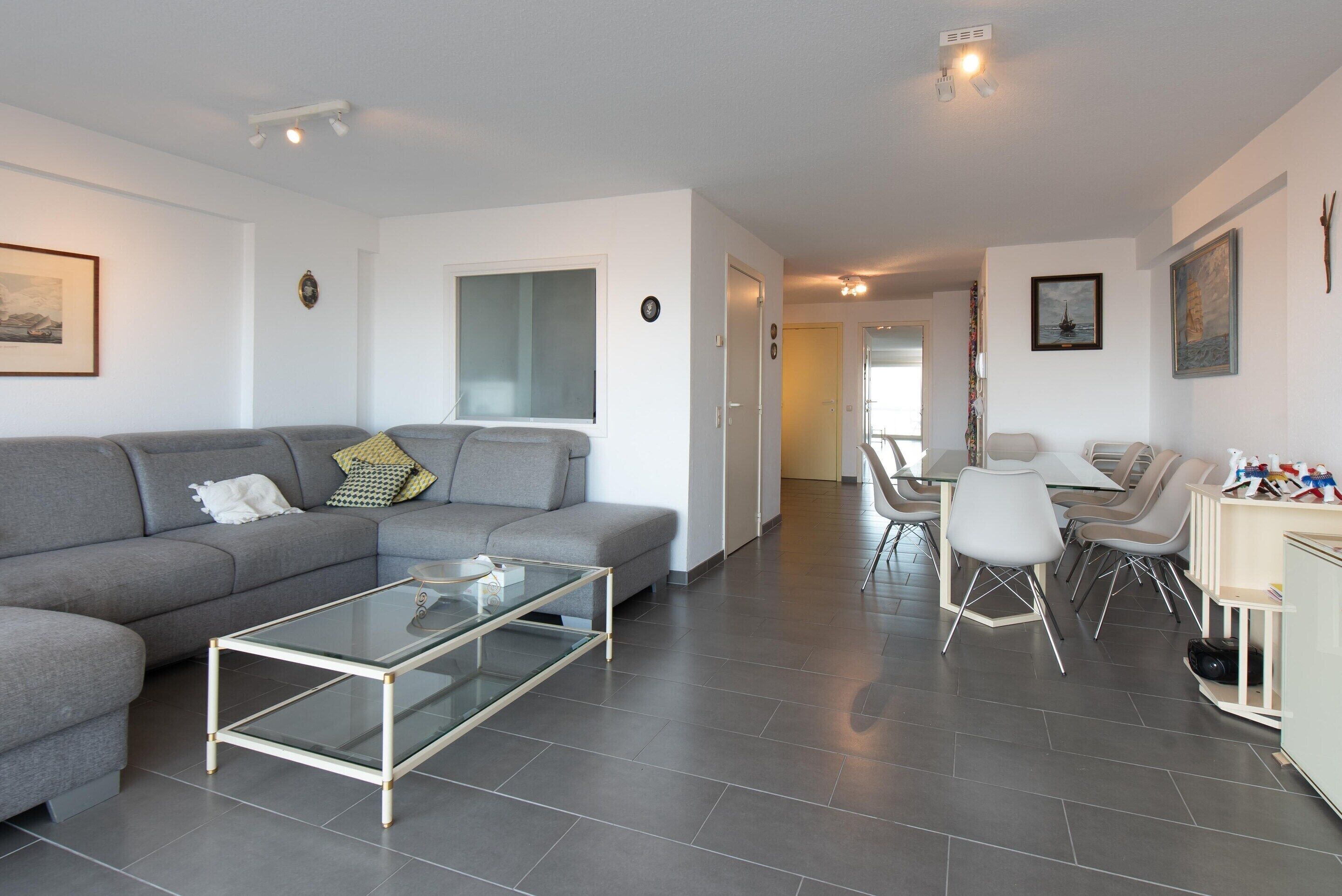 A modern living room with a grey sofa, a glass coffee table, and a dining area with white chairs and a table.