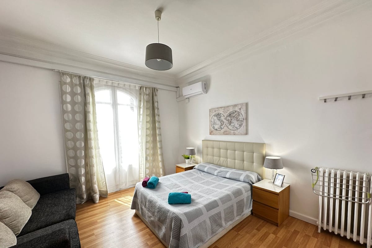 Une chambre à coucher comprenant un lit, une table de chevet, une commode, un radiateur et une fenêtre avec des rideaux.
