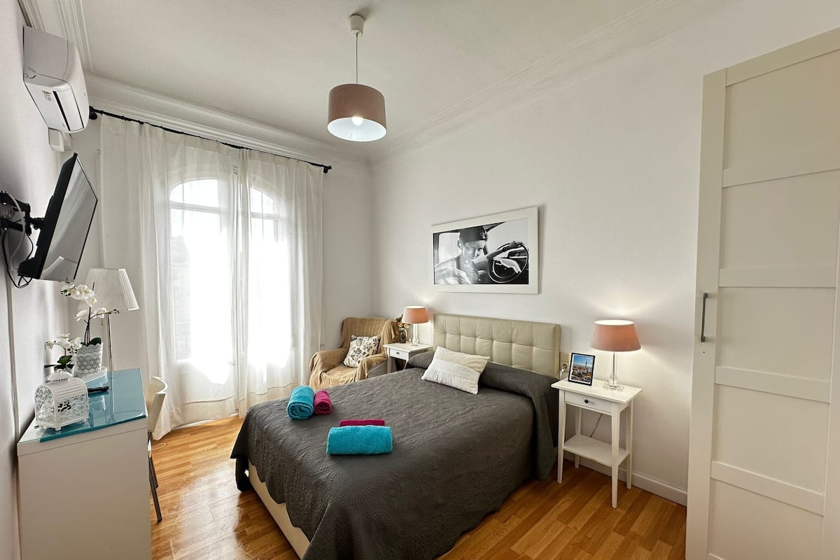 Une chambre à coucher avec un lit, des tables de chevet, une lampe, un tableau au mur et une fenêtre avec des rideaux.
