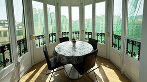 Une pièce baignée de lumière, avec une table ronde et des chaises, de grandes fenêtres ornées de balcons en fer forgé noir, et une vue sur les bâtiments environnants.