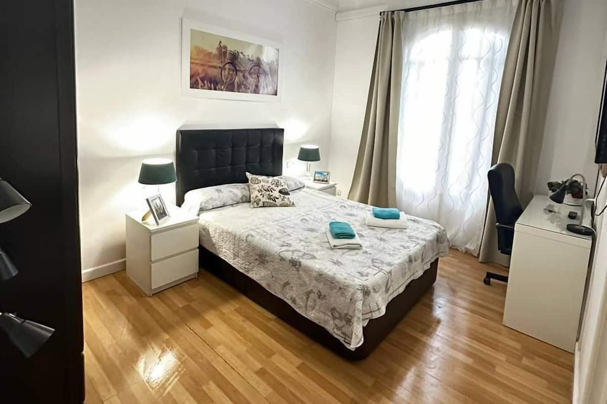 Une chambre à coucher comprenant un lit, des tables de chevet, un bureau, une chaise, une armoire, un tableau au mur et un luminaire au plafond.