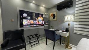 Et moderne hotelværelse med et stort fladskærms-tv, der viser en film. Der er et skrivebord med stol, et lille bord og en lampe. Værelset har et minimalistisk design med grå vægge og et hvidt gulv.