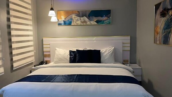 A hotel room with a bed, a nightstand, a painting above the bed, and a window with blinds.