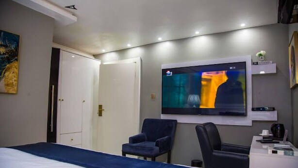 A hotel room with a bed, two chairs, a desk with a computer, and a TV mounted on the wall. - Republican City Apartment (Awka)