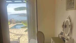 A room with a view of a beach and pool, a chair, a vanity with a mirror, and a bed.